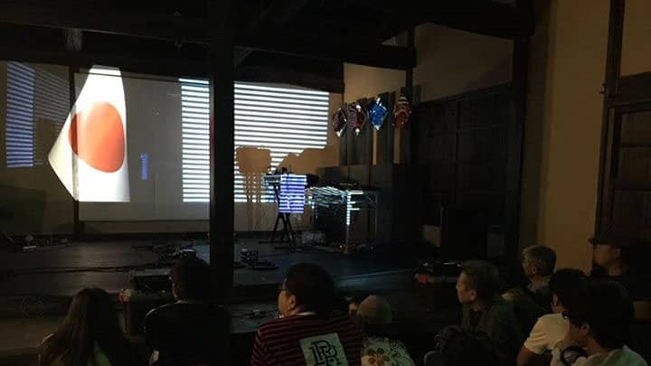 古いお屋敷で行われているアートイベントを観る観客の情景。横長の細い線と日本の国旗の映像が映されている。